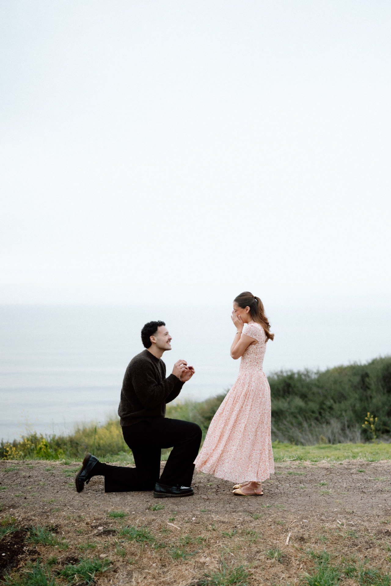 A man kneeling with a ring as his partner reacts on a coastal bluff.