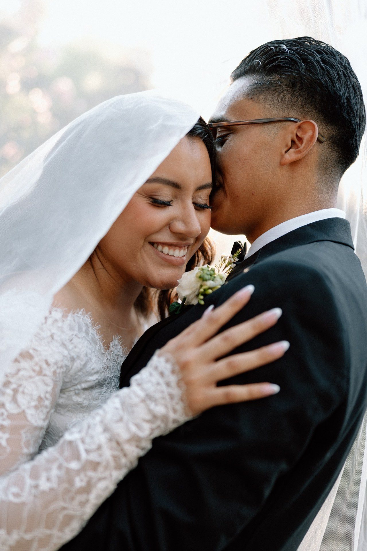 Groom kissing the bride’s forehead beneath a sunlit veil.
