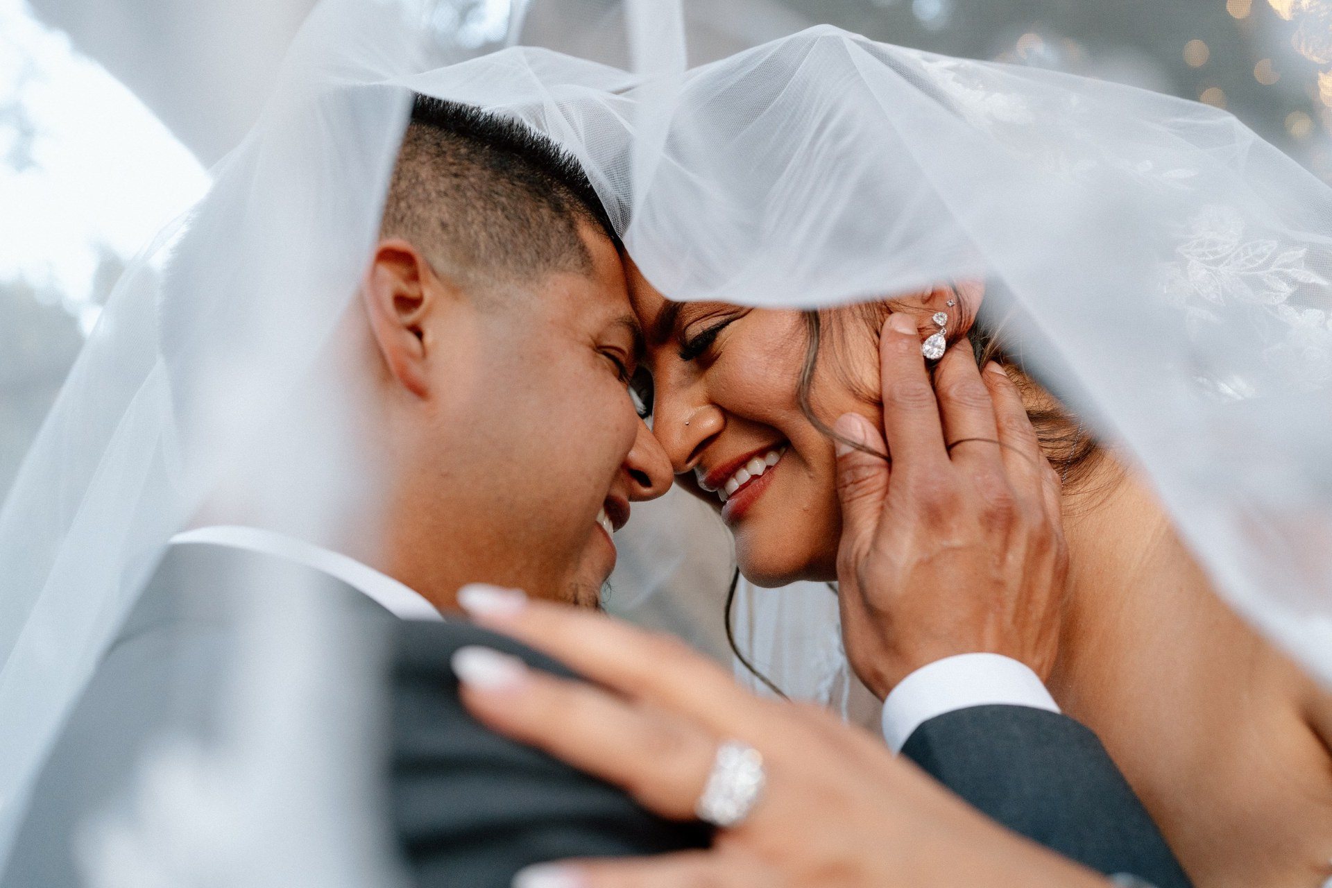 Couple smiling forehead to forehead beneath a wedding veil.