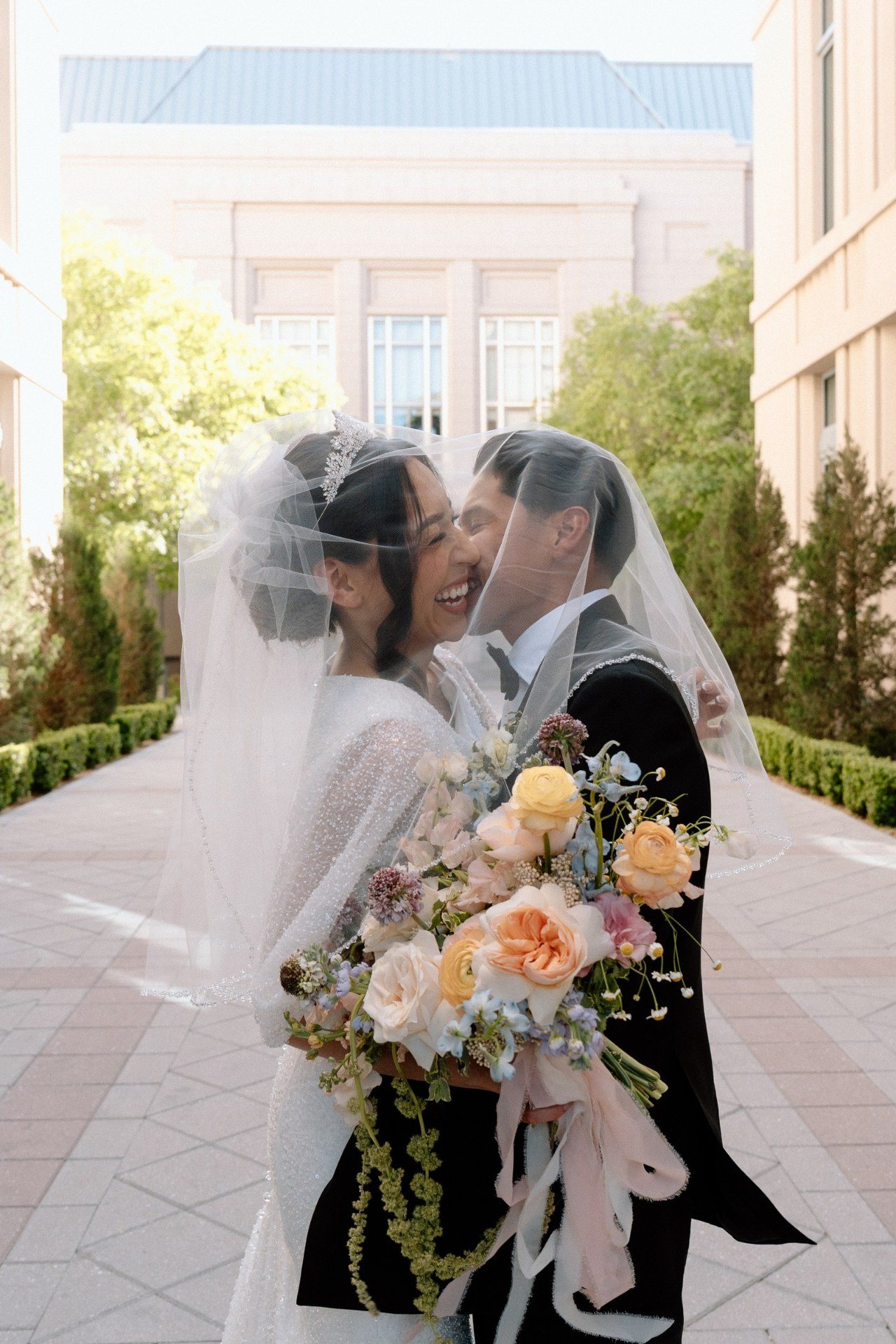 Newlyweds smiling together under a veil with a pastel bouquet.