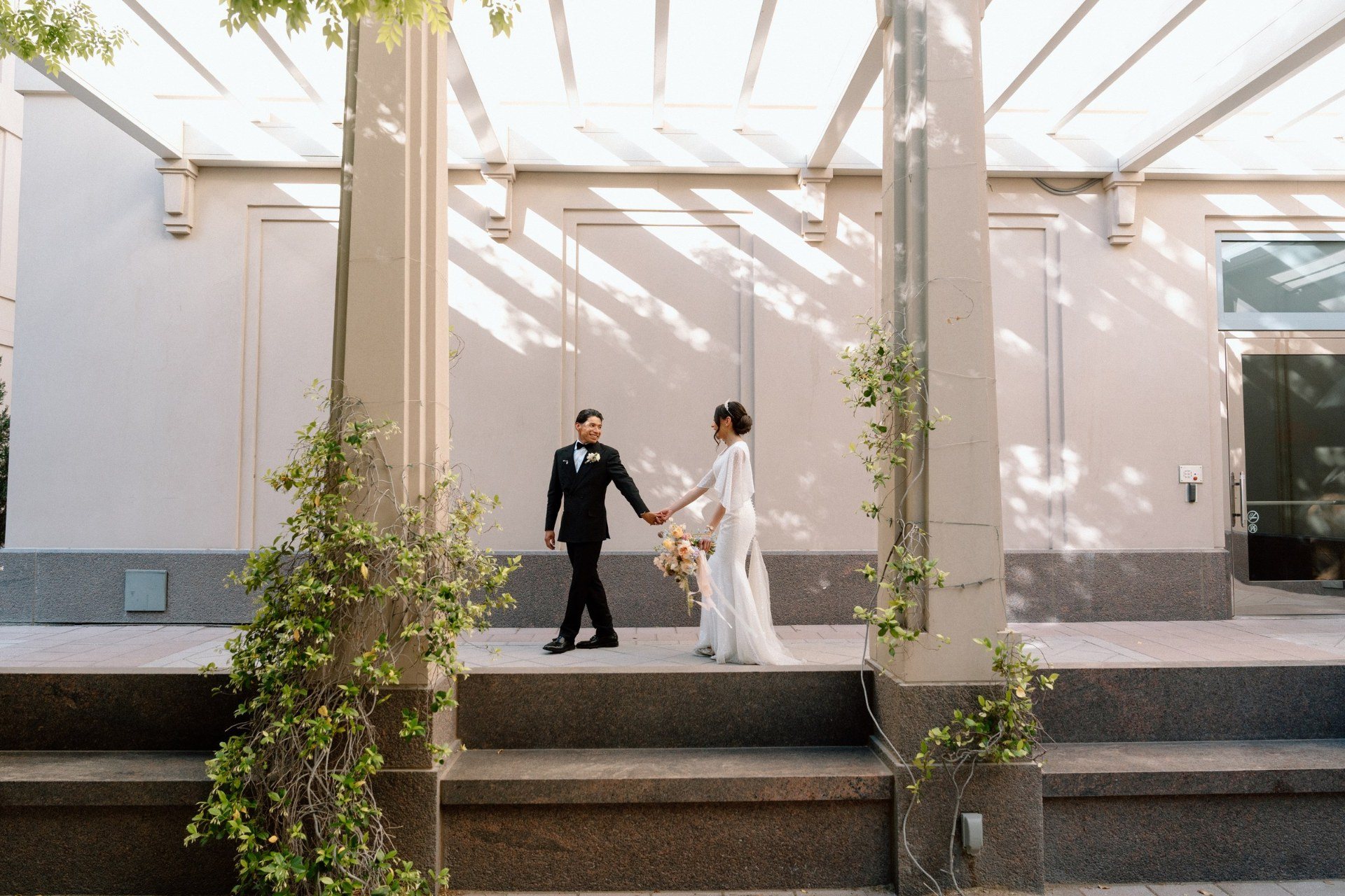 Newlyweds holding hands beneath a sunlit courtyard pergola.