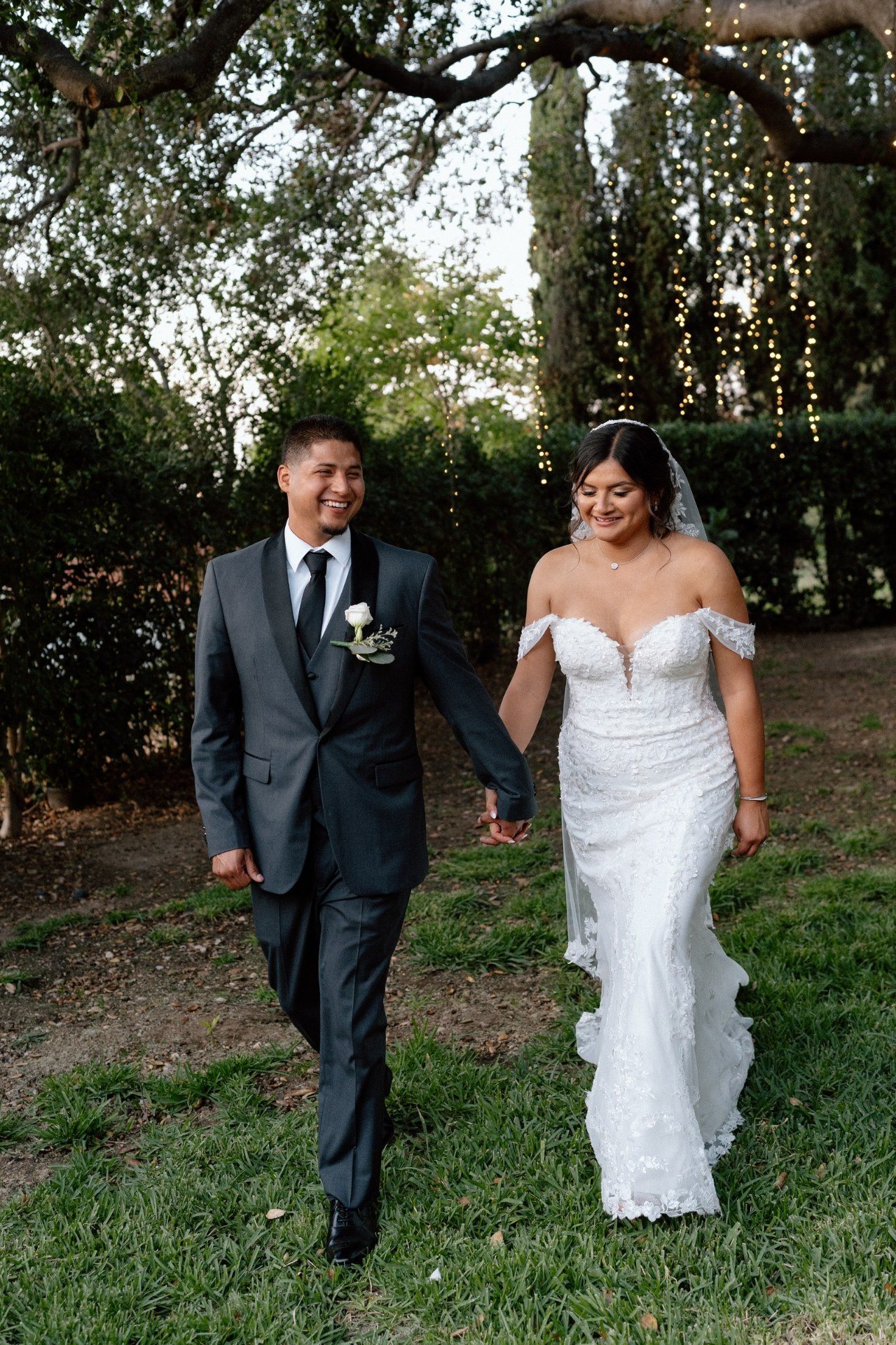 Newlyweds walking hand in hand beneath trees and string lights.