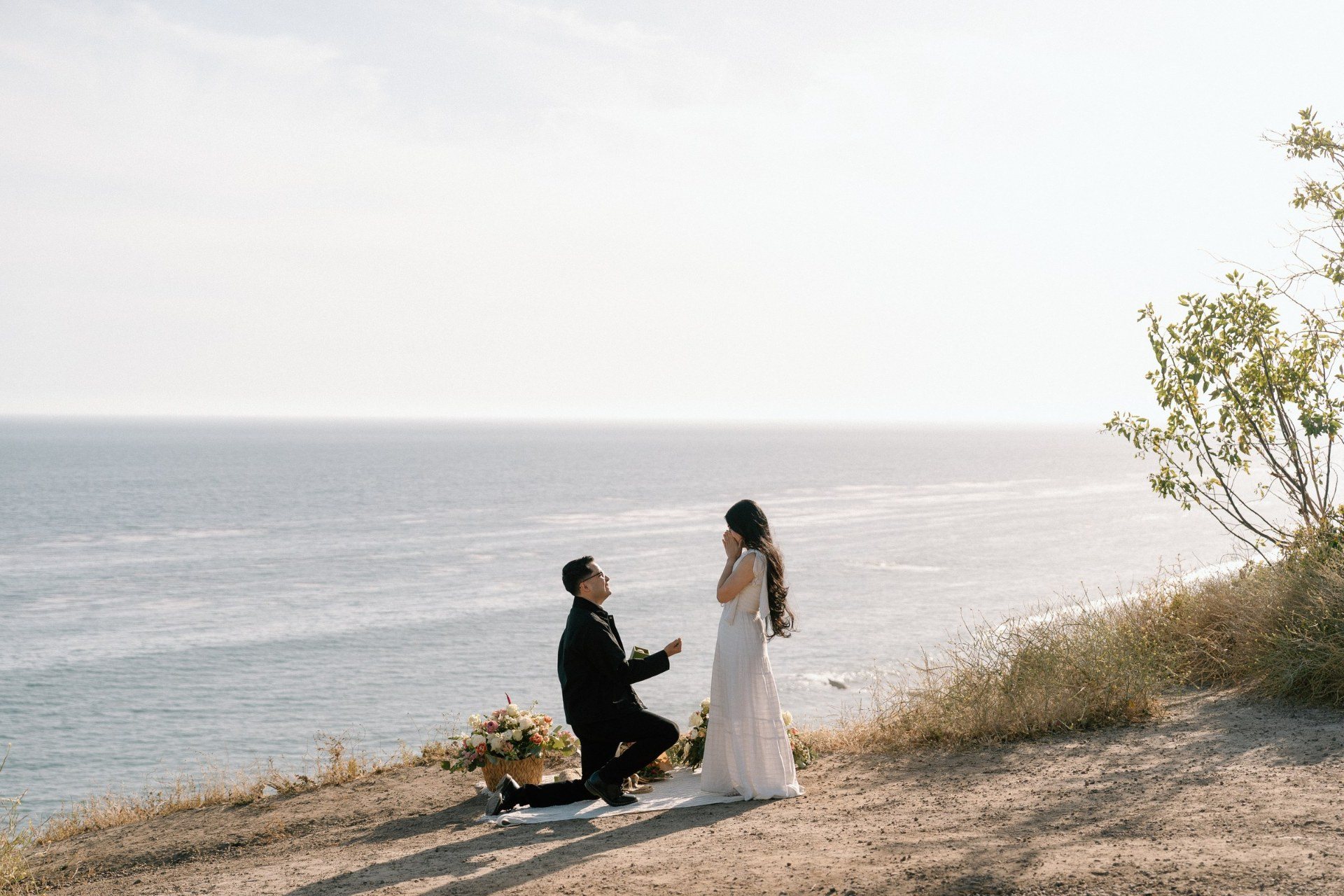 A coastal proposal with a partner kneeling beside flowers above the ocean.